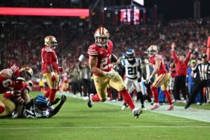 The cover for this NFL picks week 13 article shows Christian McCaffrey #23 of the San Francisco 49ers running into the end zone for a touchdown against the Carolina Panthers.