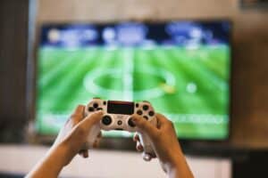 Person holding a PlayStation controller while playing a soccer video game on a large TV screen.
