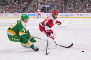 The cover for this NHL parlay picks today article shows Tyler Pitlick #19 of the Minnesota Wild defending K'Andre Miller #19 of the Carolina Hurricanes.