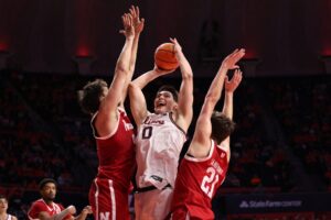 The cover for this college basketball picks today article shows David Mirkovic #0 of the Illinois Fighting Illini being guarded by Pryce Sandfort #21 of the Nebraska Cornhuskers and Berke Buyuktuncel #9 of the Nebraska Cornhuskers.