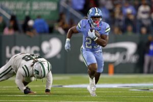 The cover for this college football player props bowls article shows Running back Javin Gordon #23 of the Tulane Green Wave running past defensive lineman Xavier Miles #58 of the Charlotte 49ers.