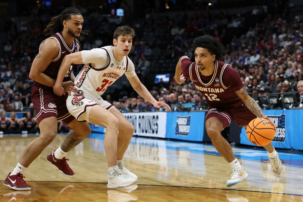 The cover for this college basketball picks today article shows Brandon Whitney #12 of the Montana Grizzlies driving to the basket past Jack Janicki #33 of the Wisconsin Badgers and Joe Pridgen #11 during the first half in the first round of the NCAA Men's Basketball Tournament.